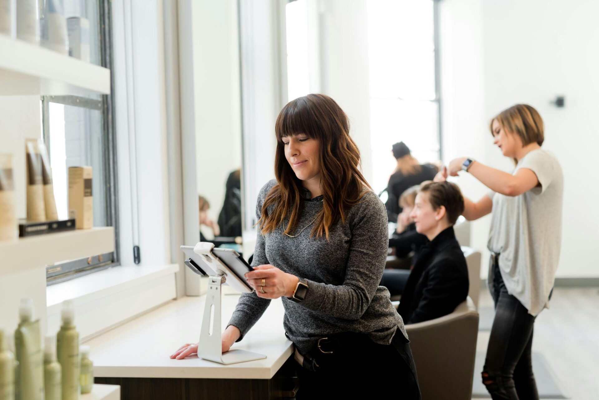 Woman standing at a salon reception desk using a tablet POS system while hairstylists work with clients in the background